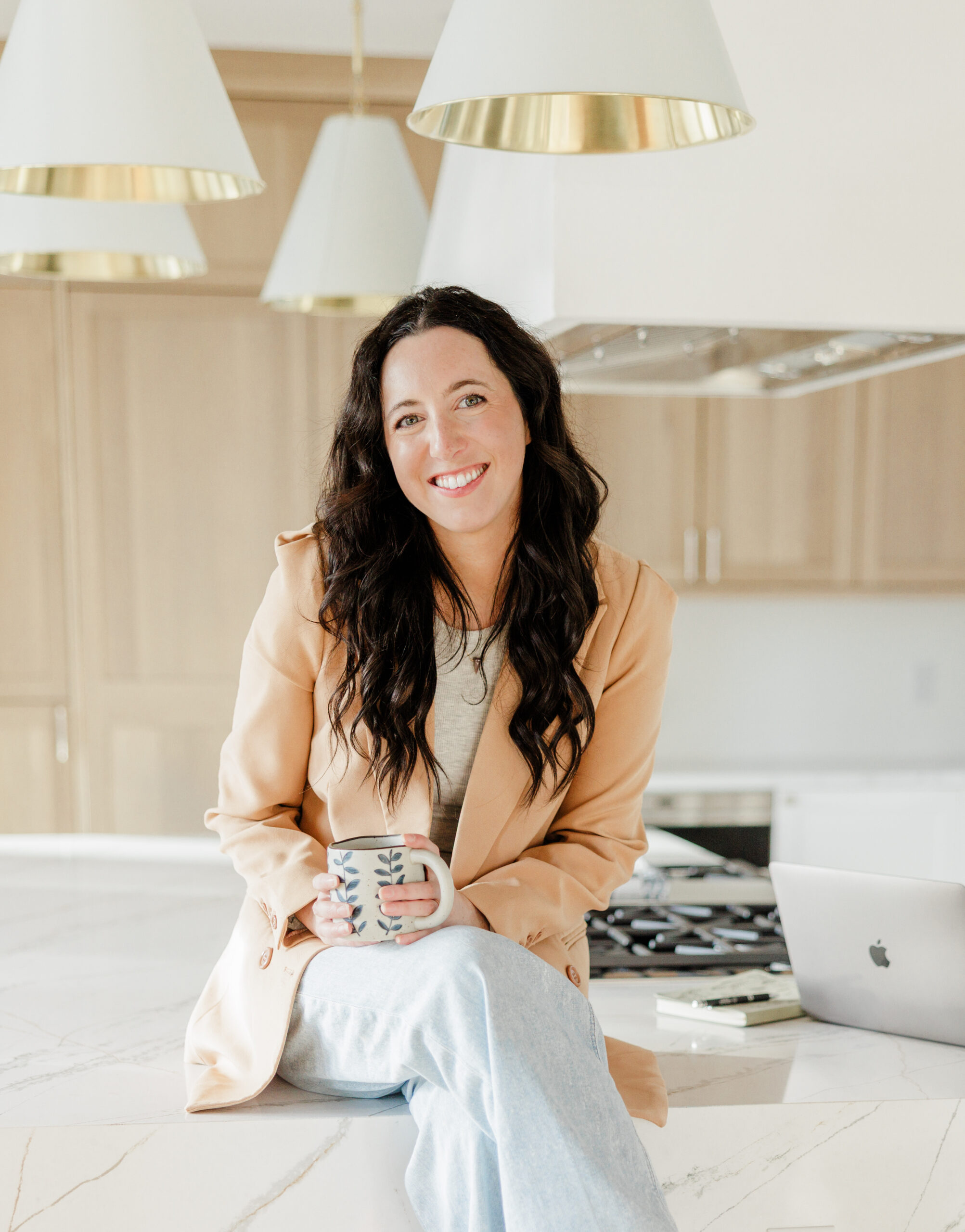 Rachel of Rachel Levin Photography sitting on a counter in a tan blazer and jeans and holding a coffee mug