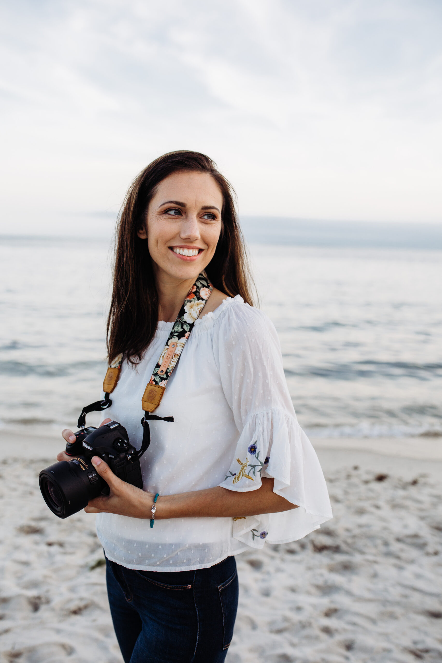 Dacia of Dacia Vu Photography  standing on the beach in a white shirt and jeans holding a camera