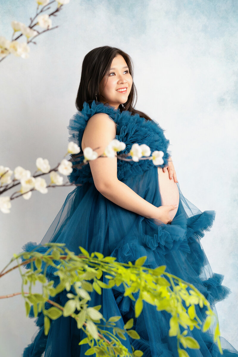 mom to be in blue tulle maternity gown standing in front of a blue and white cloudy backdrop with florals in the foreground during a session with North Austin maternity photographer Naomi & Co Photography