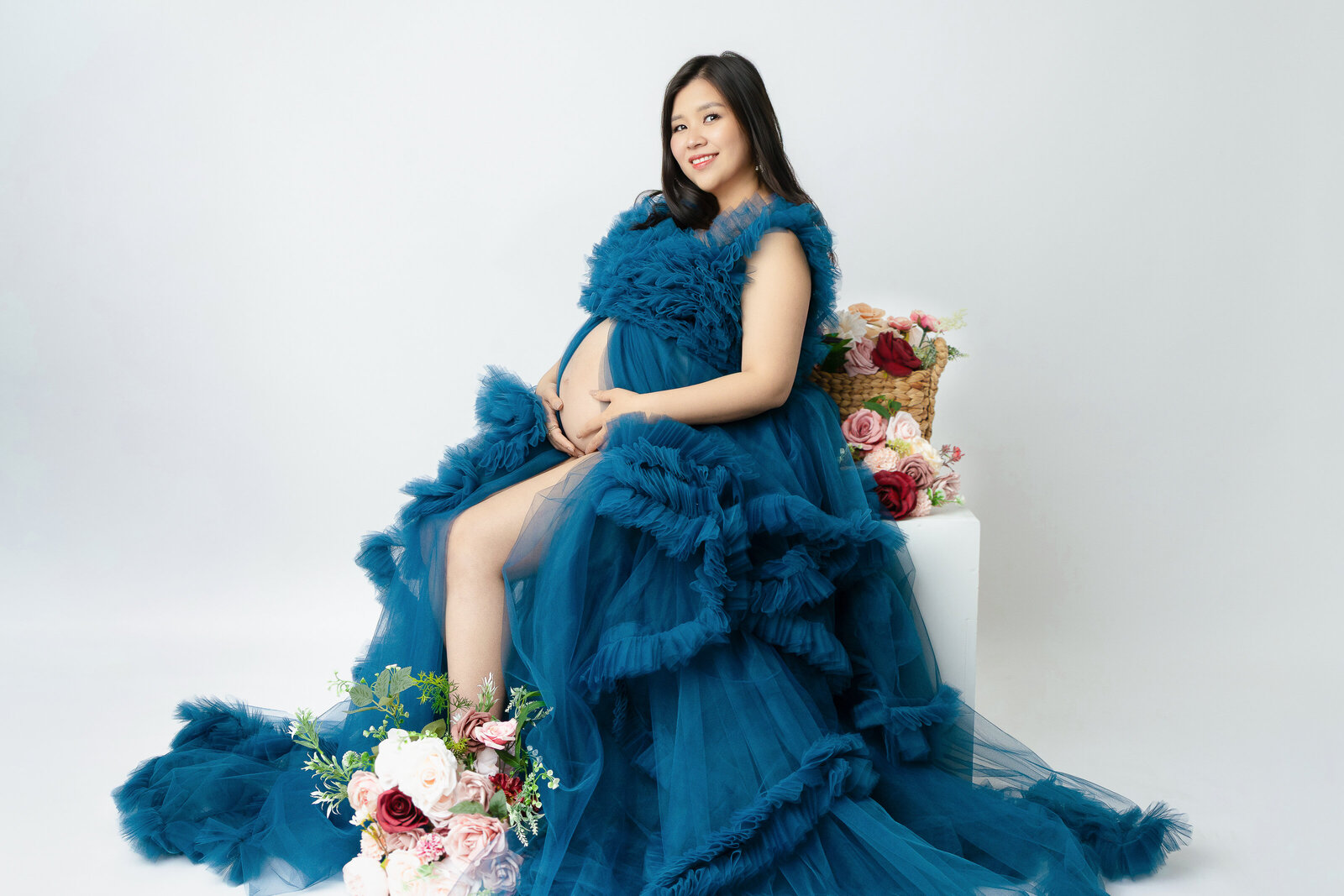 pregnant woman in blue tulle maternity gown sitting on a white cube surrounded by flowers during a session with North Austin maternity photographer Naomi & Co Photography